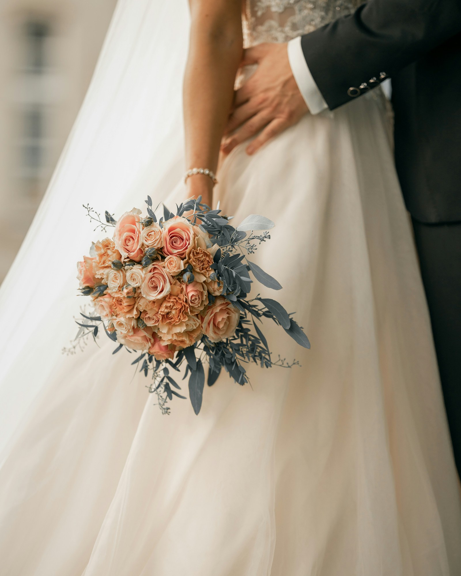 A blush rose and carnation bridal bouquet with dusty eucalyptus foliage, held against an ivory tulle gown.