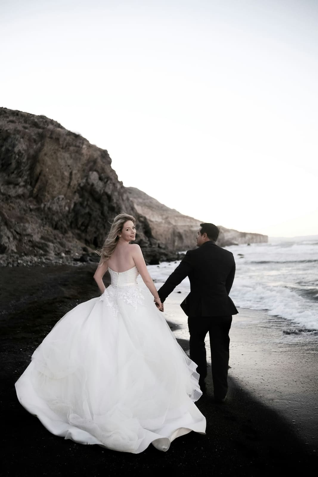 A couple walking hand in hand along a dark-sand beach below the cliffs.